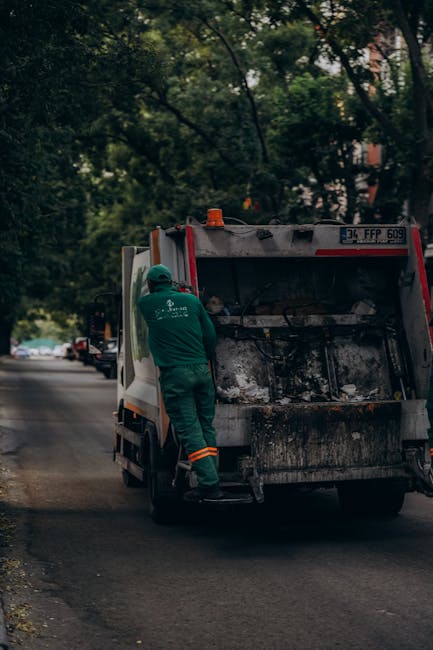 A sanitation worker dressed in a green uniform with reflective orange bands on the trousers is standing beside an open-backed rubbish collection truck parked on a quiet urban street lined with trees. 