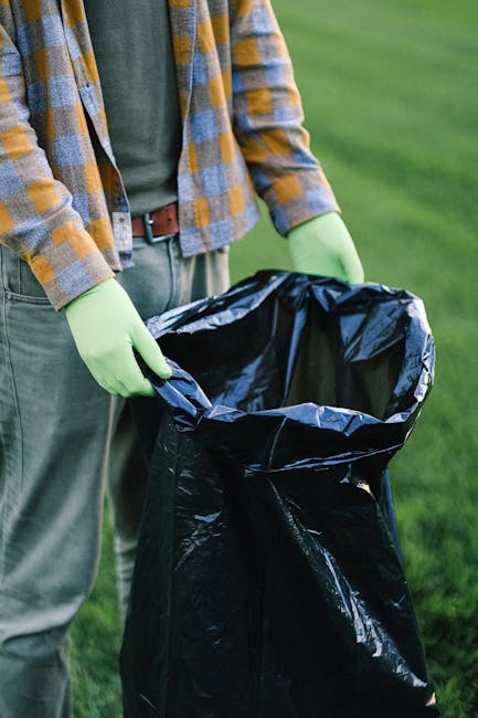 A sanitation worker dressed in a green uniform with reflective orange bands on the trousers is standing beside an open-backed rubbish collection truck parked on a quiet urban street lined with trees. 